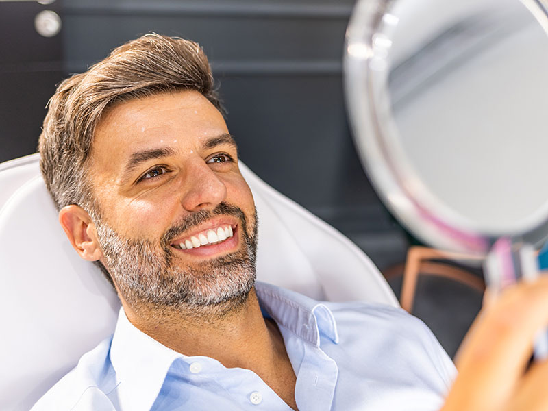 Male patient smiling while reviewing facial treatment results in a handheld mirror.